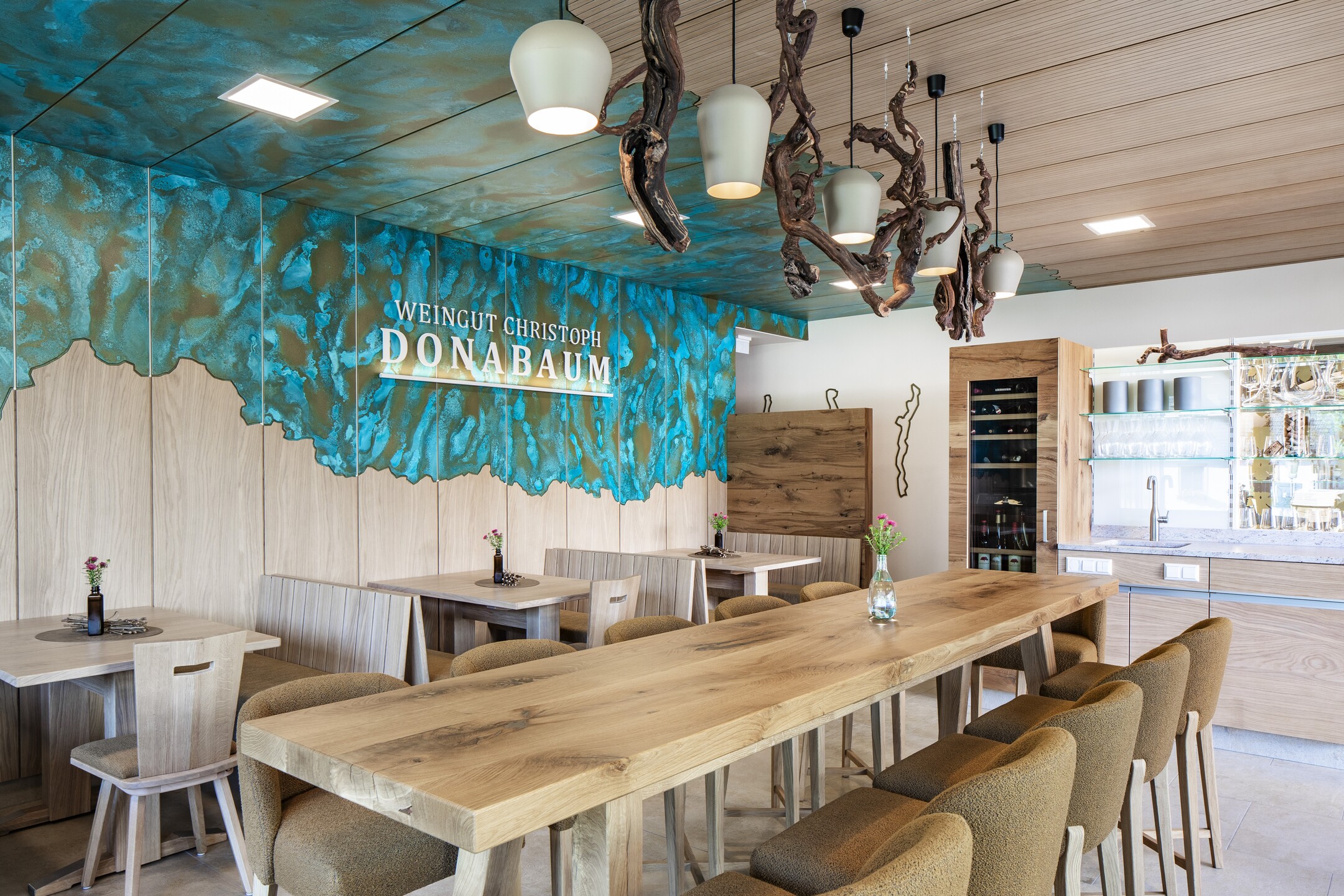 Moderne Innenraumansicht mit Logo Weingut Christoph Donabaum an der Wand, großer Holztisch mit 10 Sesseln, mehrere Esstische für Platz bis 5 Personen