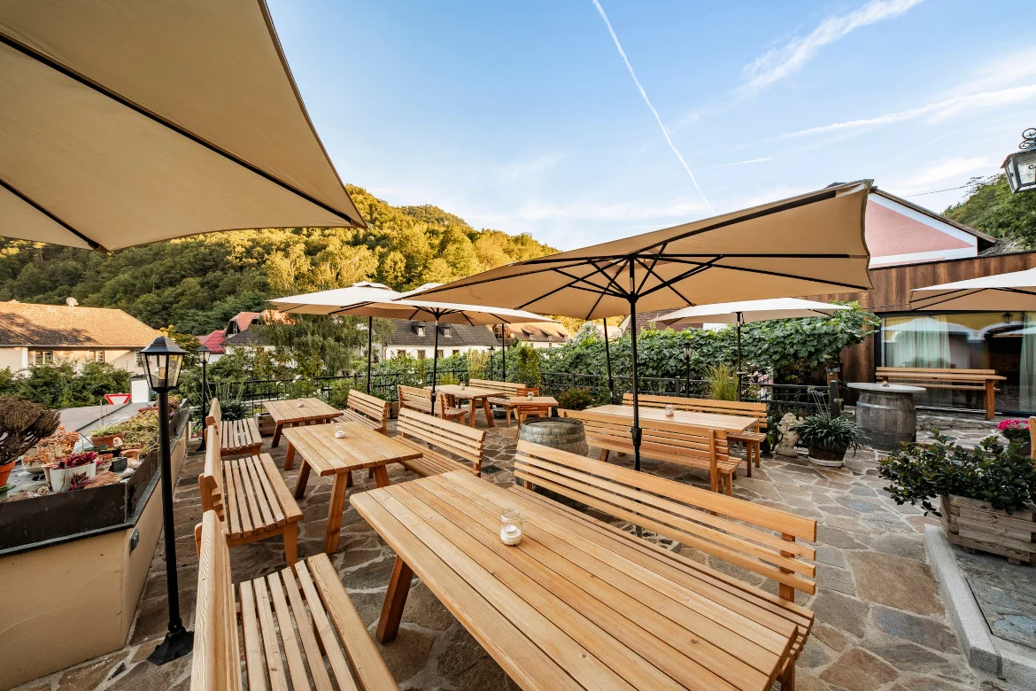 Heurigenterrasse außen mit Holztischen und Holzbänken mit Lehnen und hellbraunen Sonnenschirmen dazwischen.